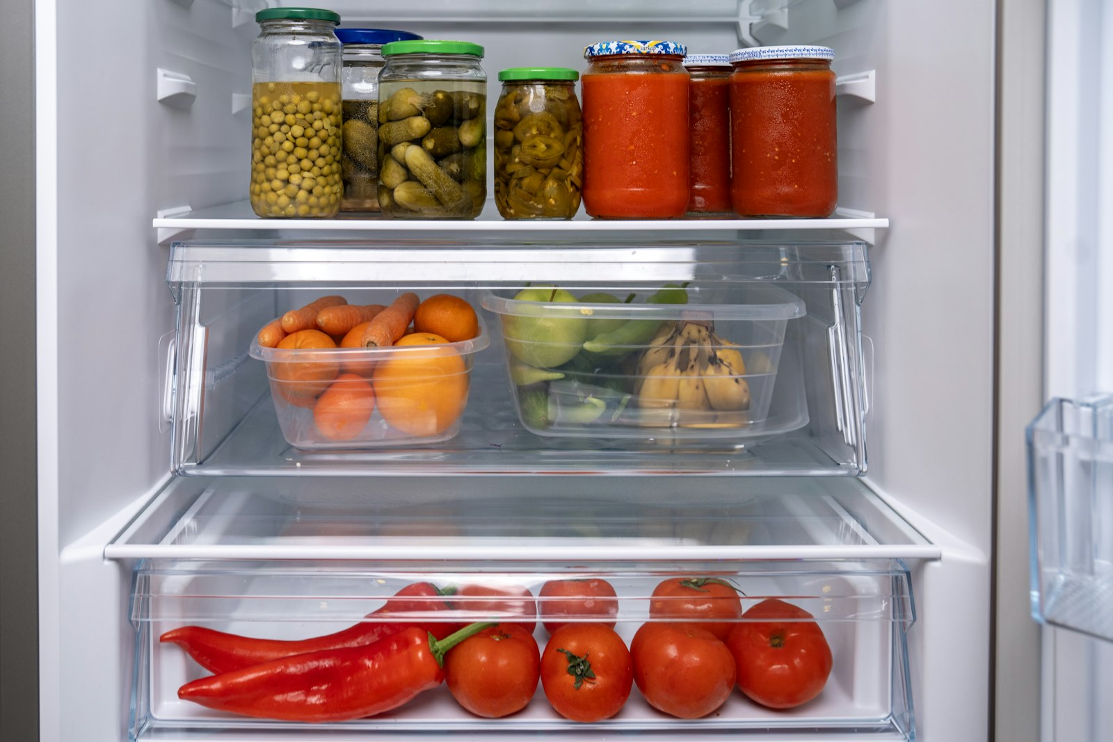 Offener Kühlschrank mit Einmachgläsern, Karotten, Orangen, Äpfeln, Bananen, Paprika und Tomaten