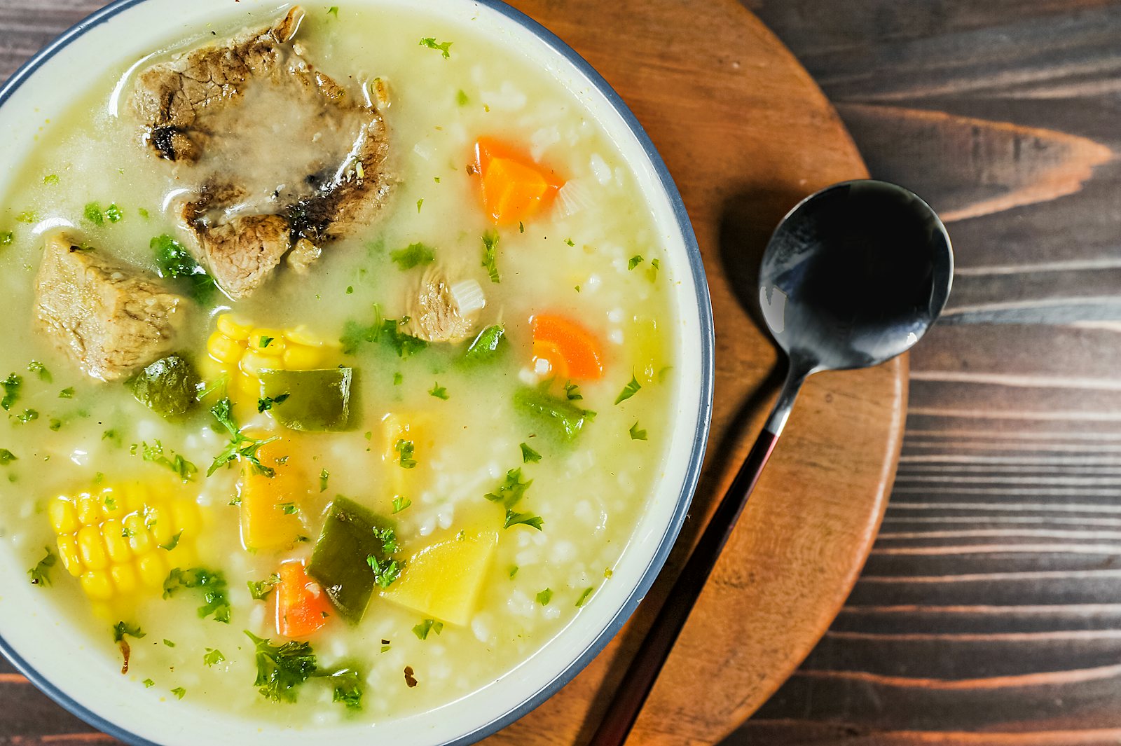 Teller mit Gemüsesuppe mit Fleisch, Mais, Karotten und Kräutern, daneben ein schwarzer Löffel auf einem Holzbrett
