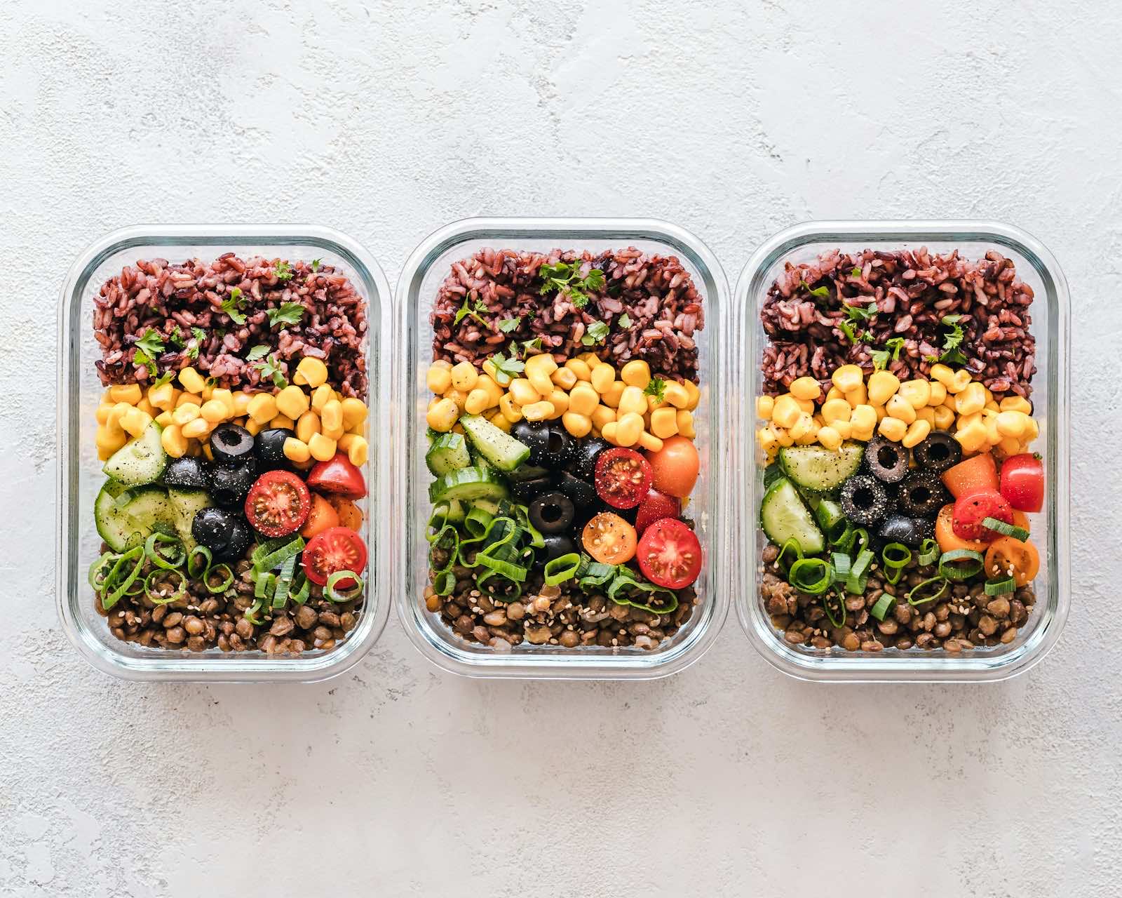 Drei Glasbehälter mit Reis, Mais, Gurke, Oliven, Tomaten und Linsen nebeneinander von oben