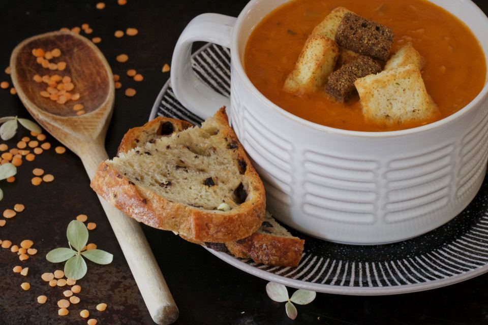 Schüssel mit roter Linsensuppe, Croûtons und Brot, daneben ein Holzlöffel mit roten Linsen
