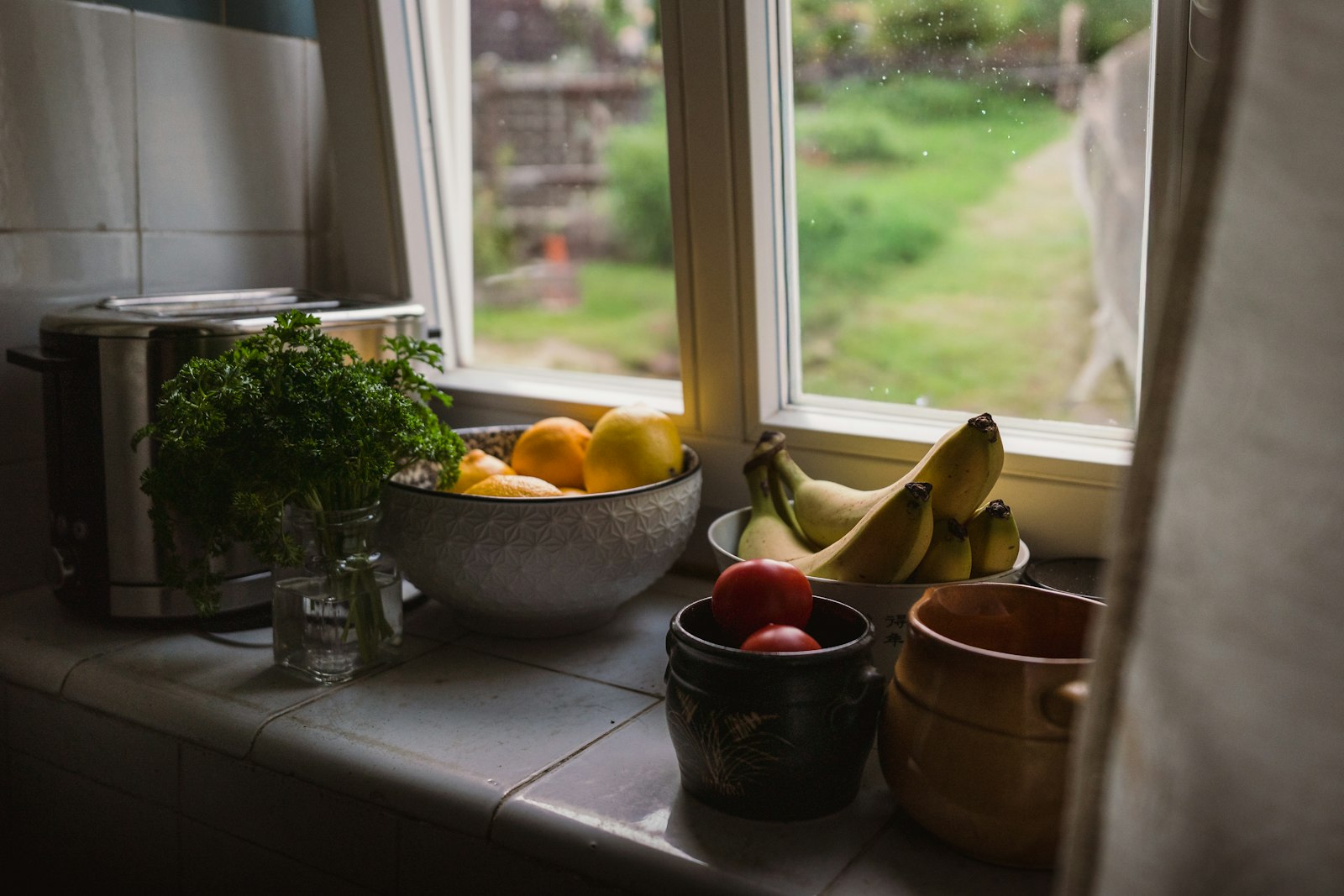 Fensterbank in einer Küche mit einer Schale Zitronen und Orangen, Bananen, Tomaten in einem Topf und einem Bund Petersilie